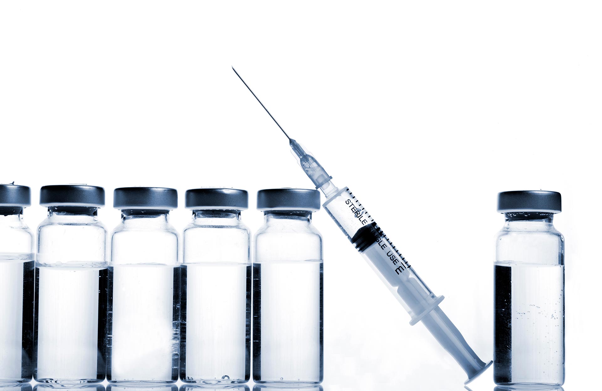 The image shows a collection of glass vials containing what appears to be medication, with a syringe placed among them, set against a backdrop of additional vials and a wall that resembles a chalkboard.
